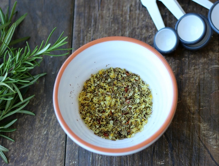 Dried herbs and spices in a white bowl with an orange rim, measuring spoons and fresh rosemary on a wooden table. 