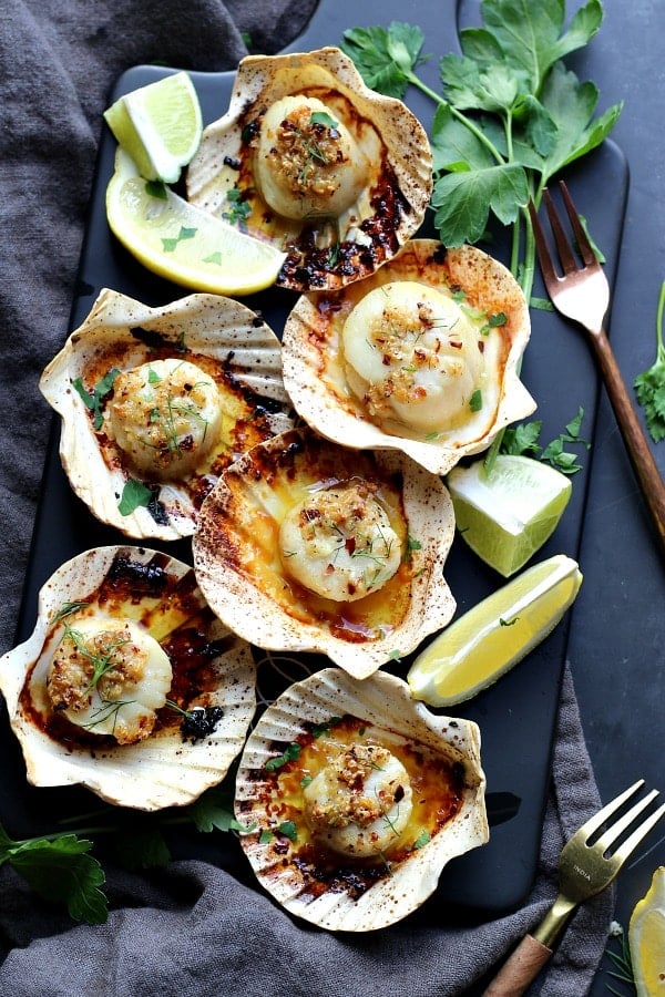 Sea scallops baked in a sea shell, seasoned with chili garlic oil. Lime slices and fresh parsley on the table. A small fork and kitchen towel on the table. 