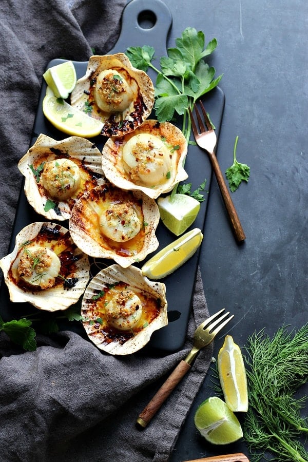 Sea scallops baked in a sea shell, seasoned with chili garlic oil. Lime slices and fresh parsley on the table. A small fork and kitchen towel on the table. 