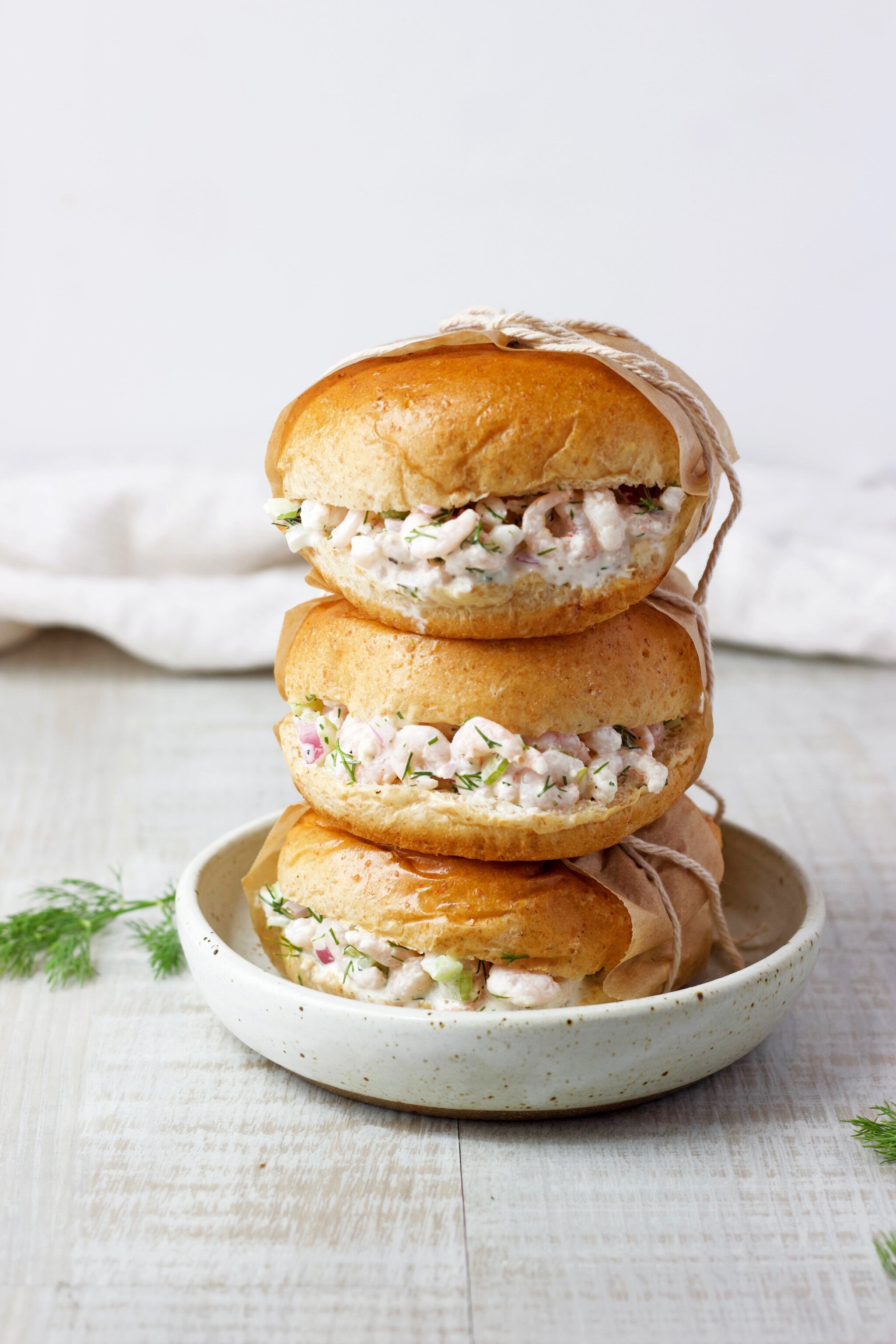 Three shrimp salad sandwiches on top of each other, sitting on a shallow bowl. Fresh dill and a towel on the table. 
