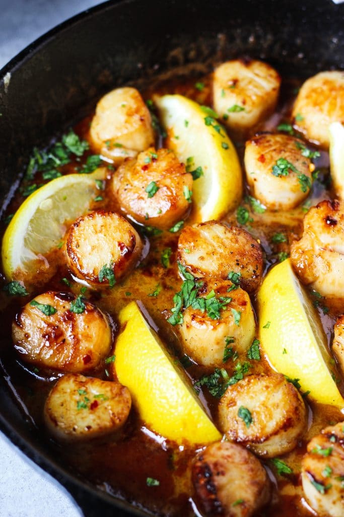 Scallops in butter and lemon in a cast iron skillet. Lemon wedges and fresh herbs on top.