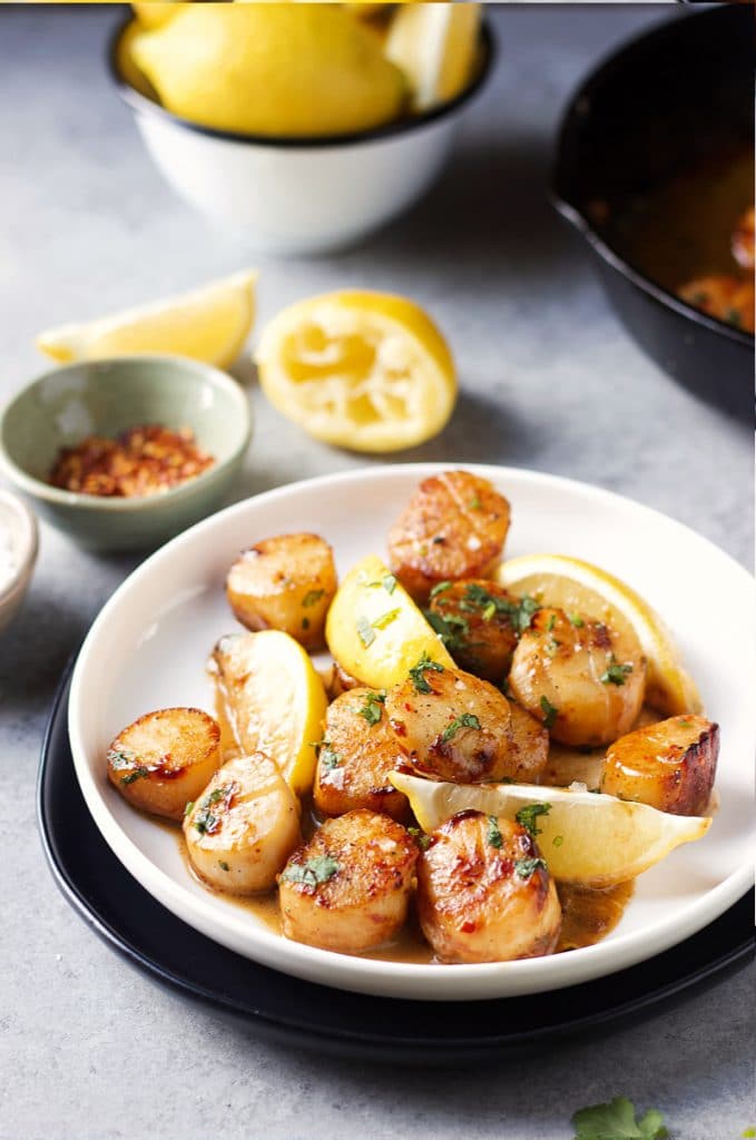 Scallops in a white round plate with lemon wedges. A pinch bowl with red pepper flakes and lemon slices on the table.