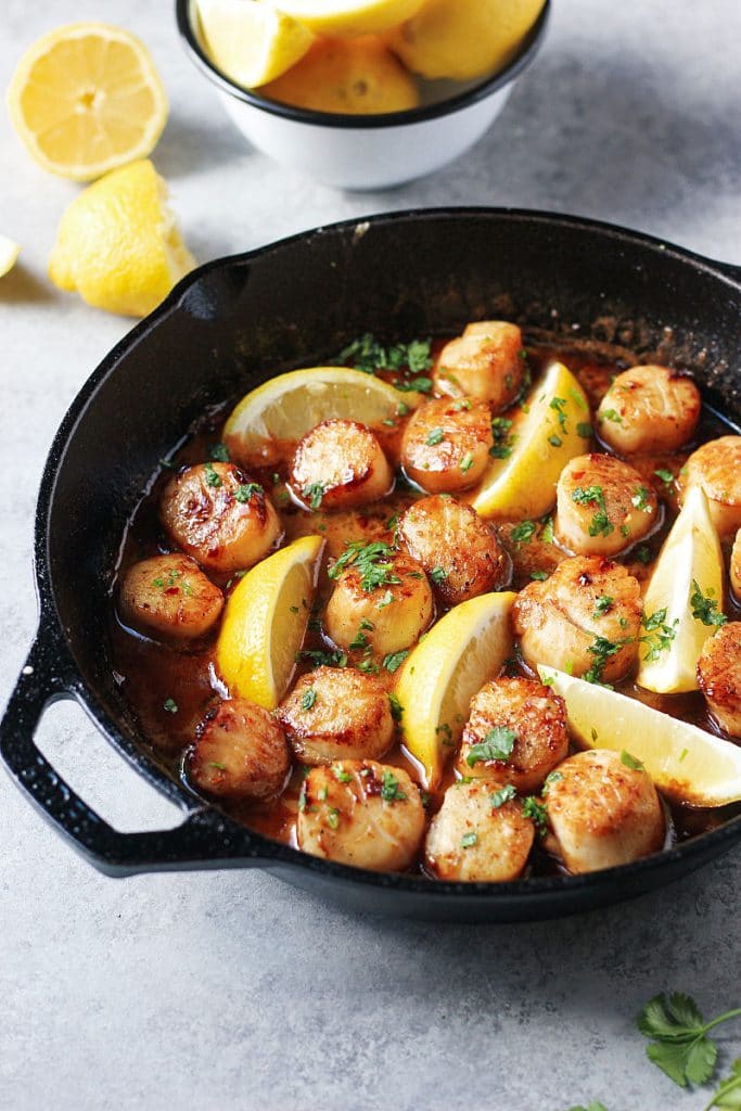 Scallops in cast iron skillet in sauce with lemon wedges and fresh herbs on top. 