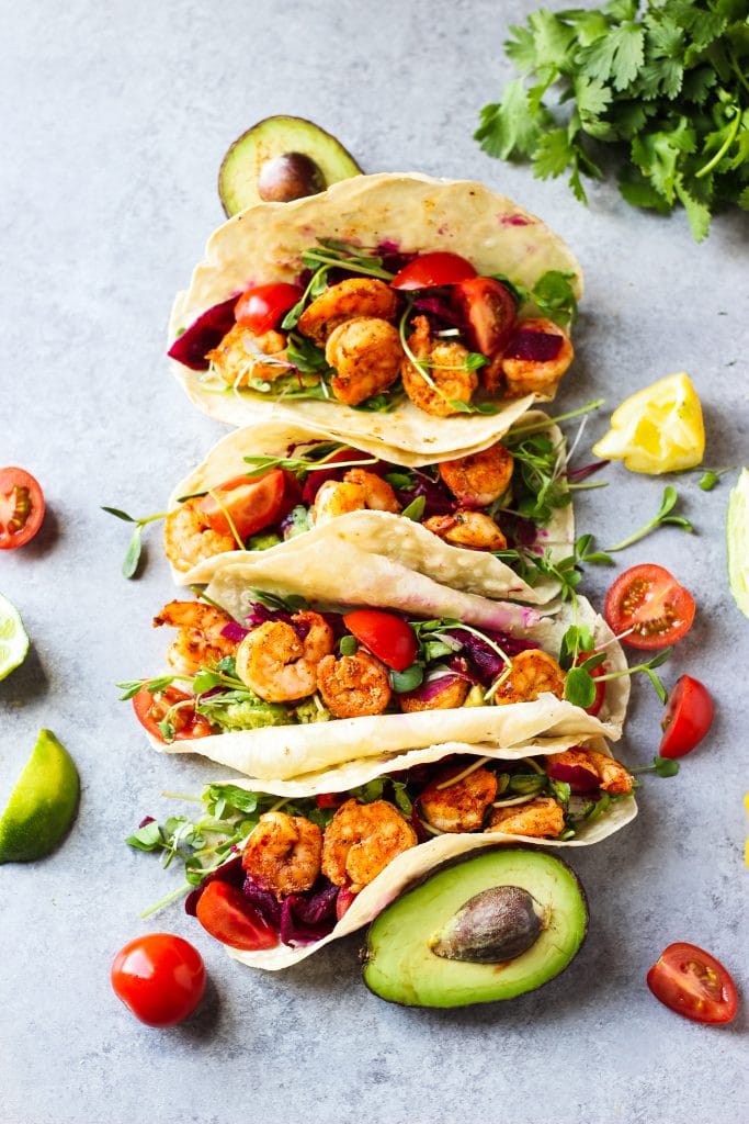 Four Shrimp Tacos arranged neatly surrounded by slices of tomatoes, lime and avocado.