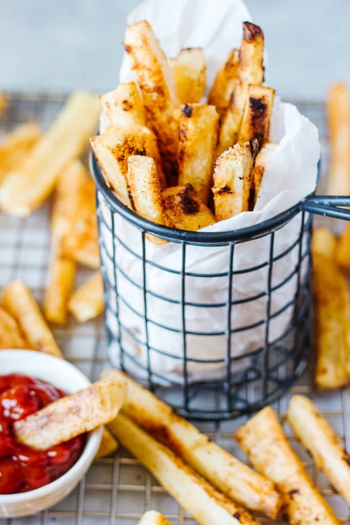 Slices of baked yuca in a cooling rack and in a  wire mesh container with a small container of ketchup and a small piece of the fries in it
