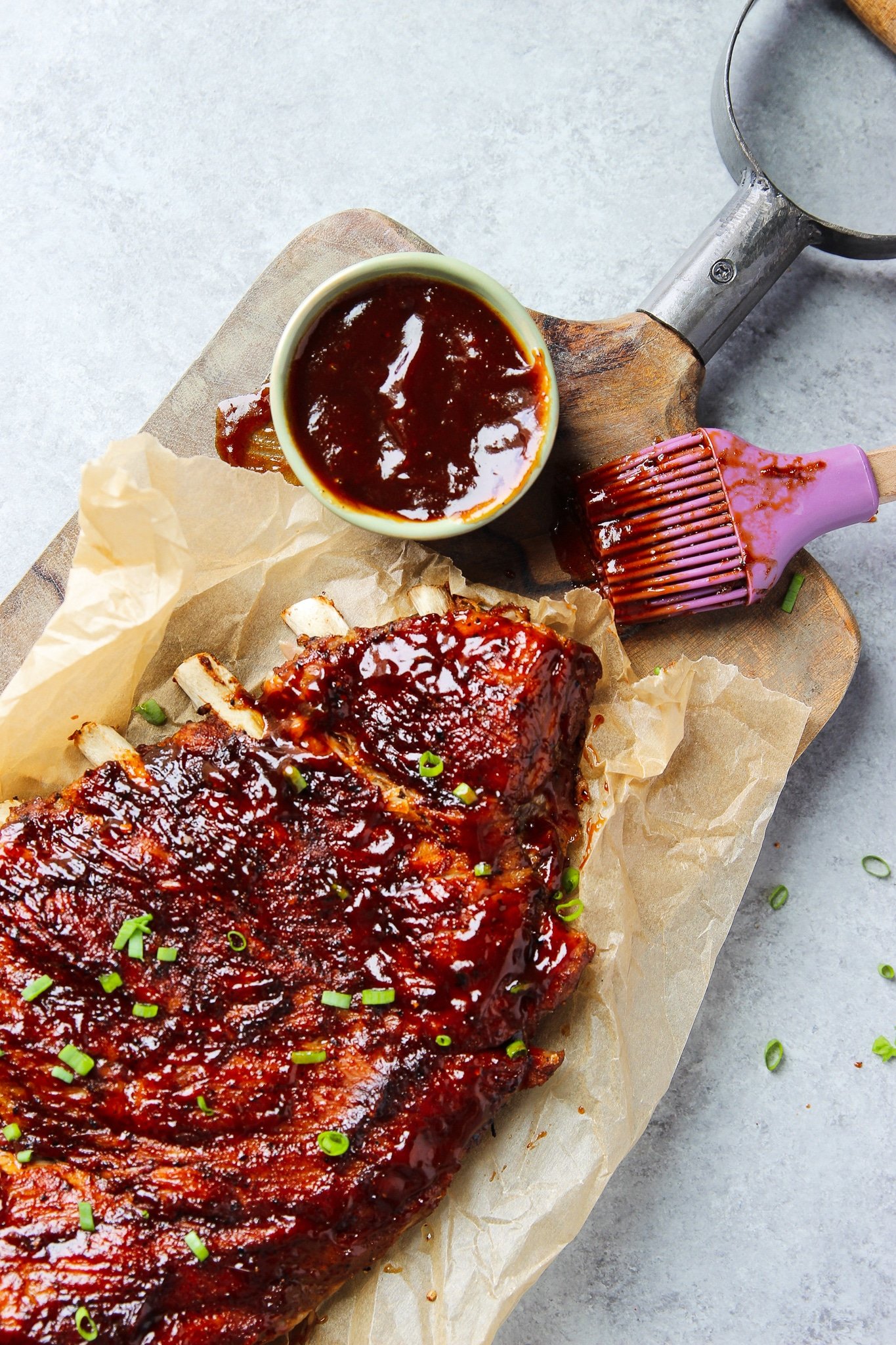 a rack of cooked bbq ribs on a parchment-lined cutting board next to a silicone brush and small bowl of bbq sauce.