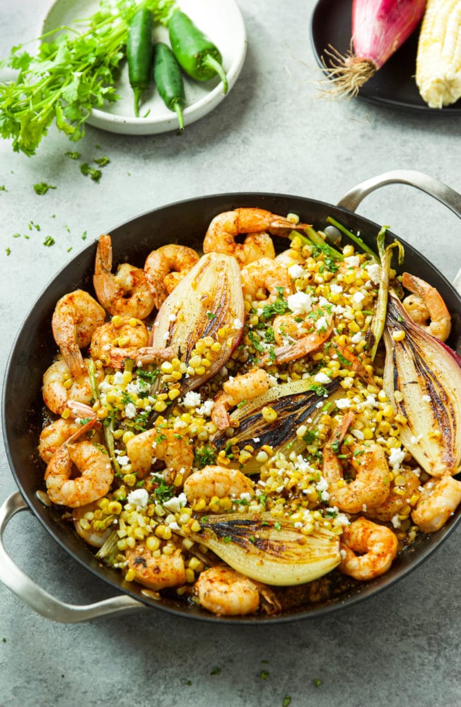 Cast iron skillet with jumbo shrimp, grilled onions, corn, feta cheese and cilantro.  A white dish with cilantro and jalapenos and a black dish with corn on the cobb and red onion.