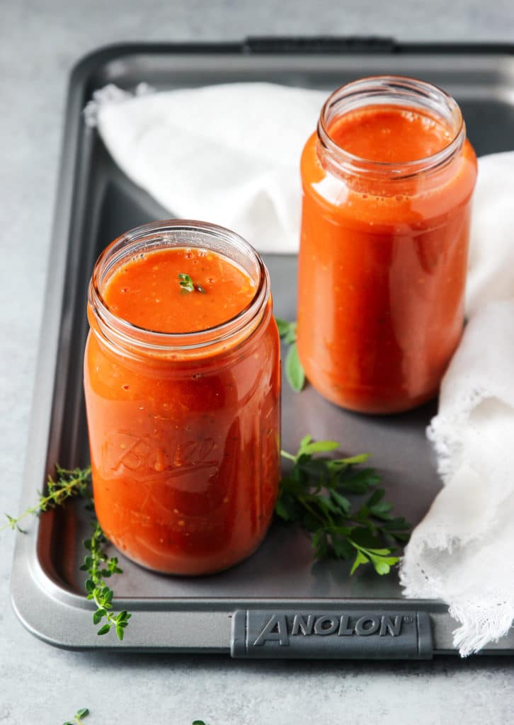 A couple of glass jars of tomato sauce with a white towel on a cookie sheet with fresh rosemary and parsley.