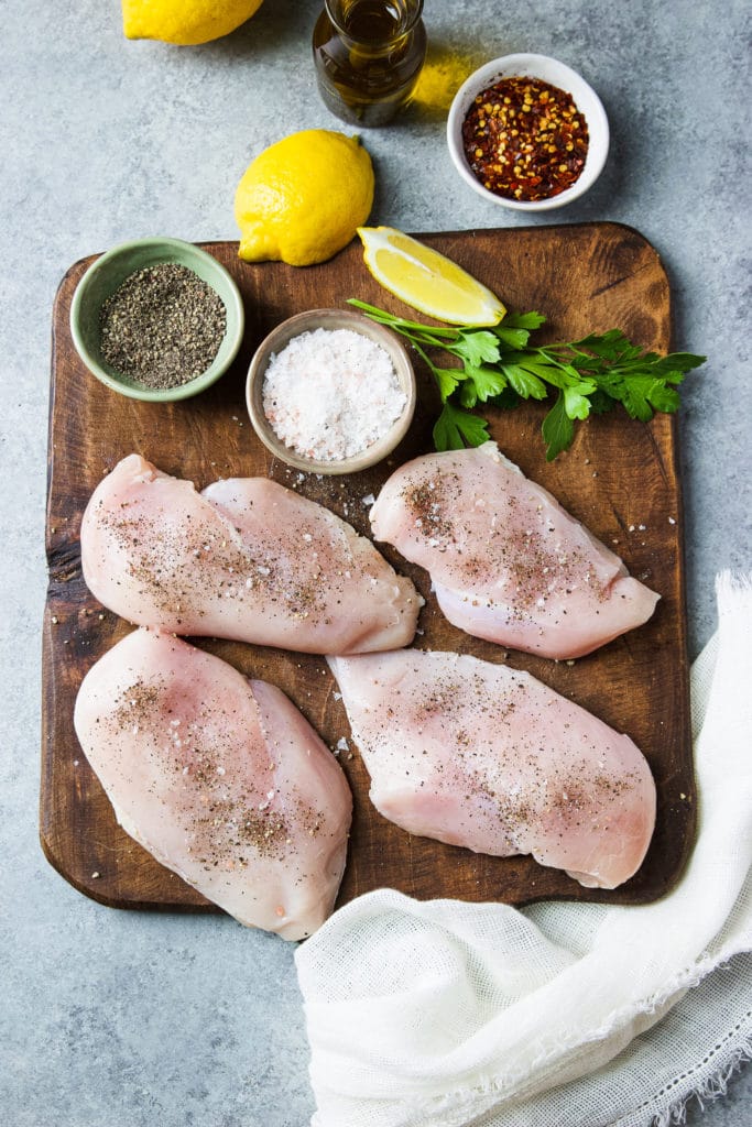 Uncooked skinless boneless chicken cutlets on a wooden chopping board surrounded by spices
