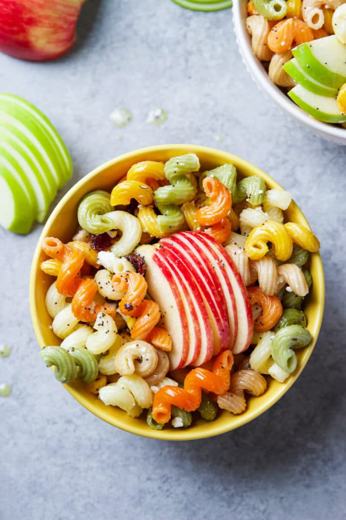 A big bowl of pasta salad with colorful noodles, sliced apples and pears with poppy seeds dressing in a yellow bowl