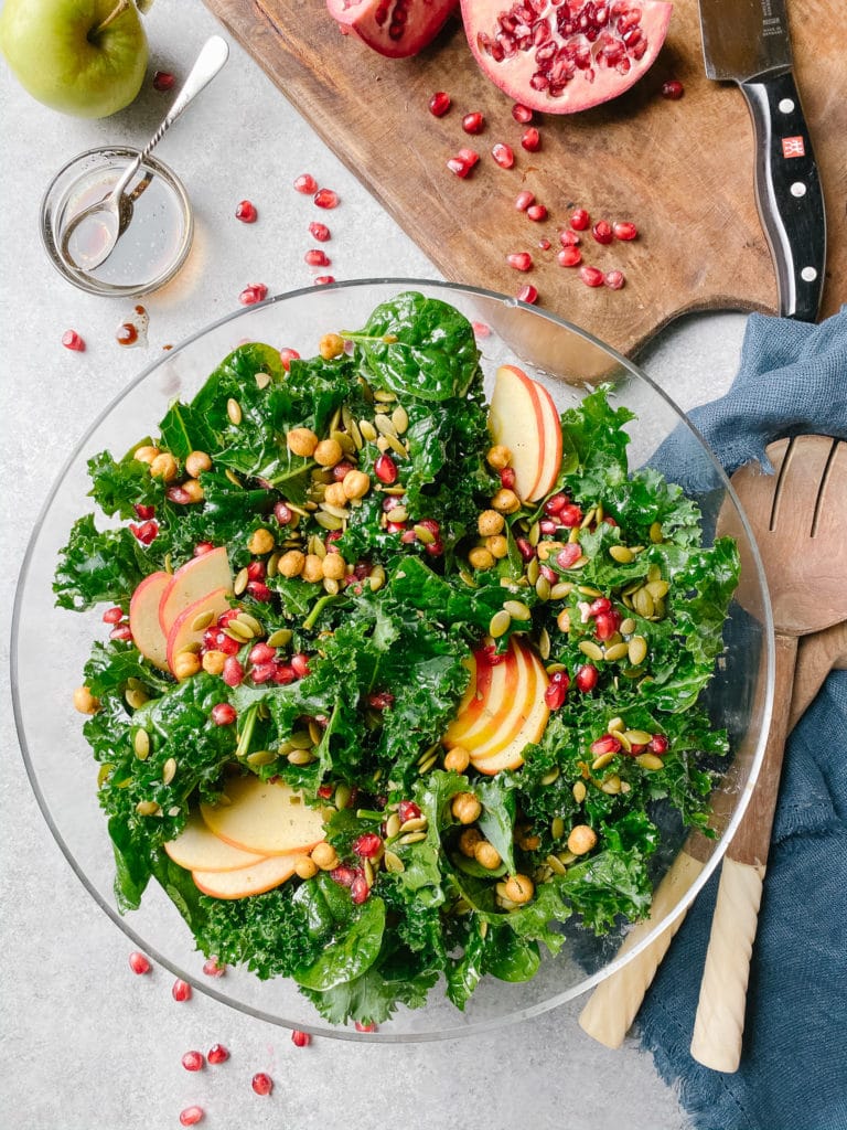 Kale, slices of apple, baby spinach, chickpeas, and pumpkin seeds in a large glass bowl with wooden spoons, dressing in a small bowl and pomegranate sliced in half on a chopping board with knife