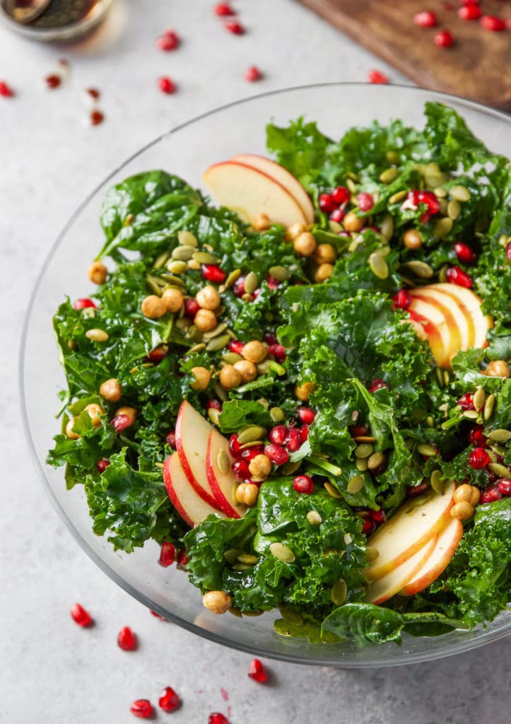 Colorful salad in a glass bowl containing kale leaves, spinach, apple slices, pomegranate seeds, sunflower seeds, and chickpeas