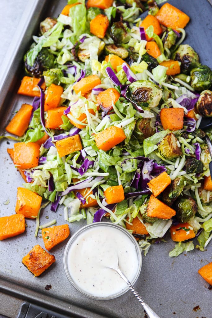 Mixed greens and roasted vegetables mixed on a sheet pan with a bowl of dressing