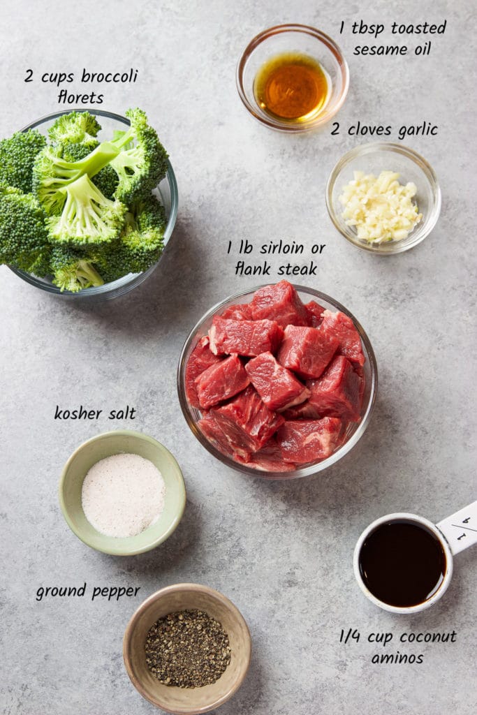 Raw ingredients consisting of broccoli florets and beef along with small glass containers of sesame oil, chopped garlic, kosher salt, ground black pepper and coconut aminos