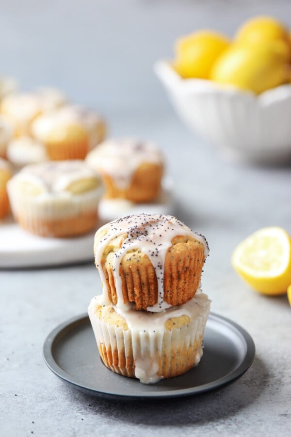 Gluten-Free Lemon Poppy Seed Muffins on top of another topped with icing and some muffins and whole lemons in the background