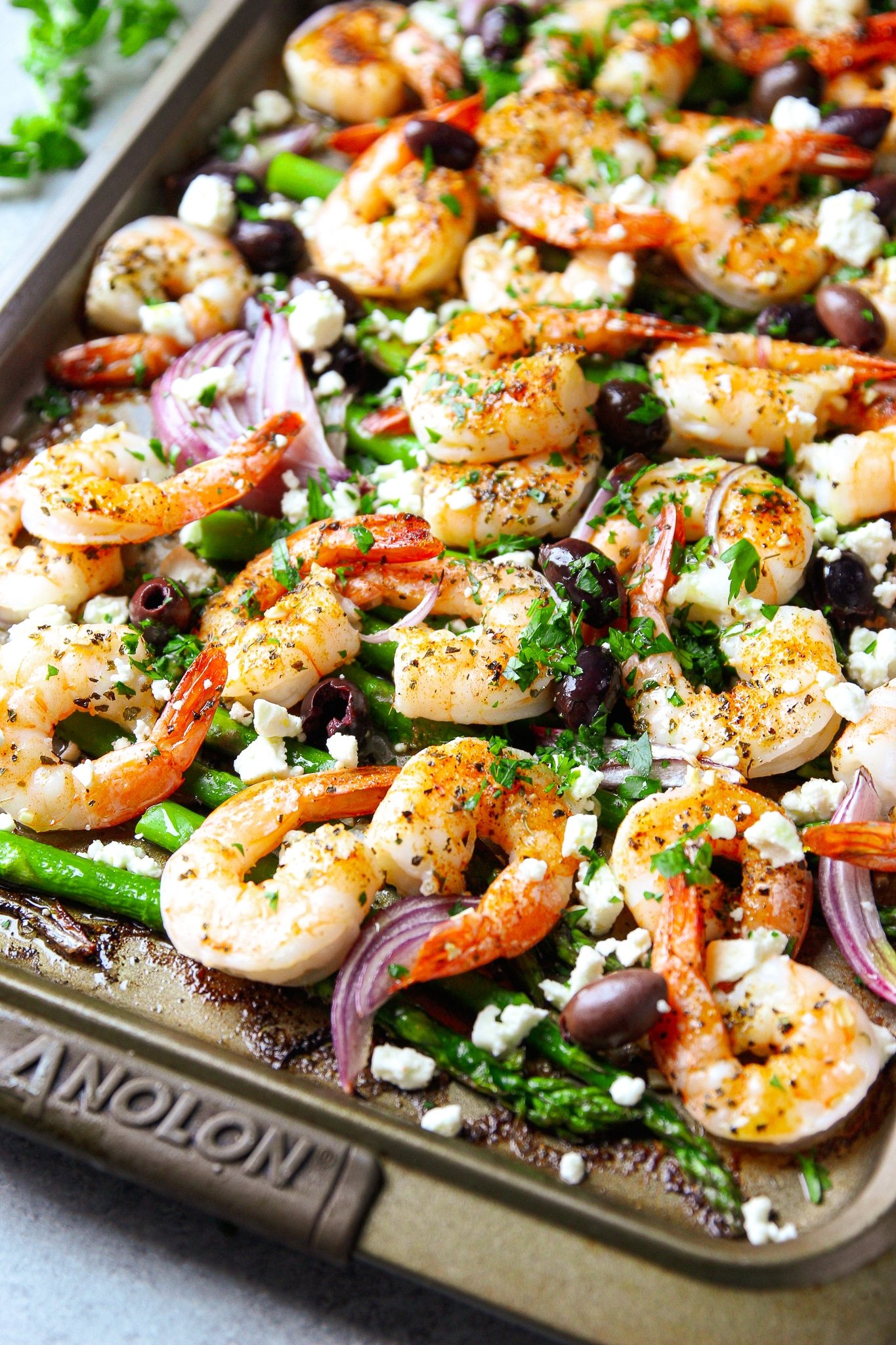 close up on roasted shrimp, red onions, and asparagus garnished with herbs and feta on a sheet pan.