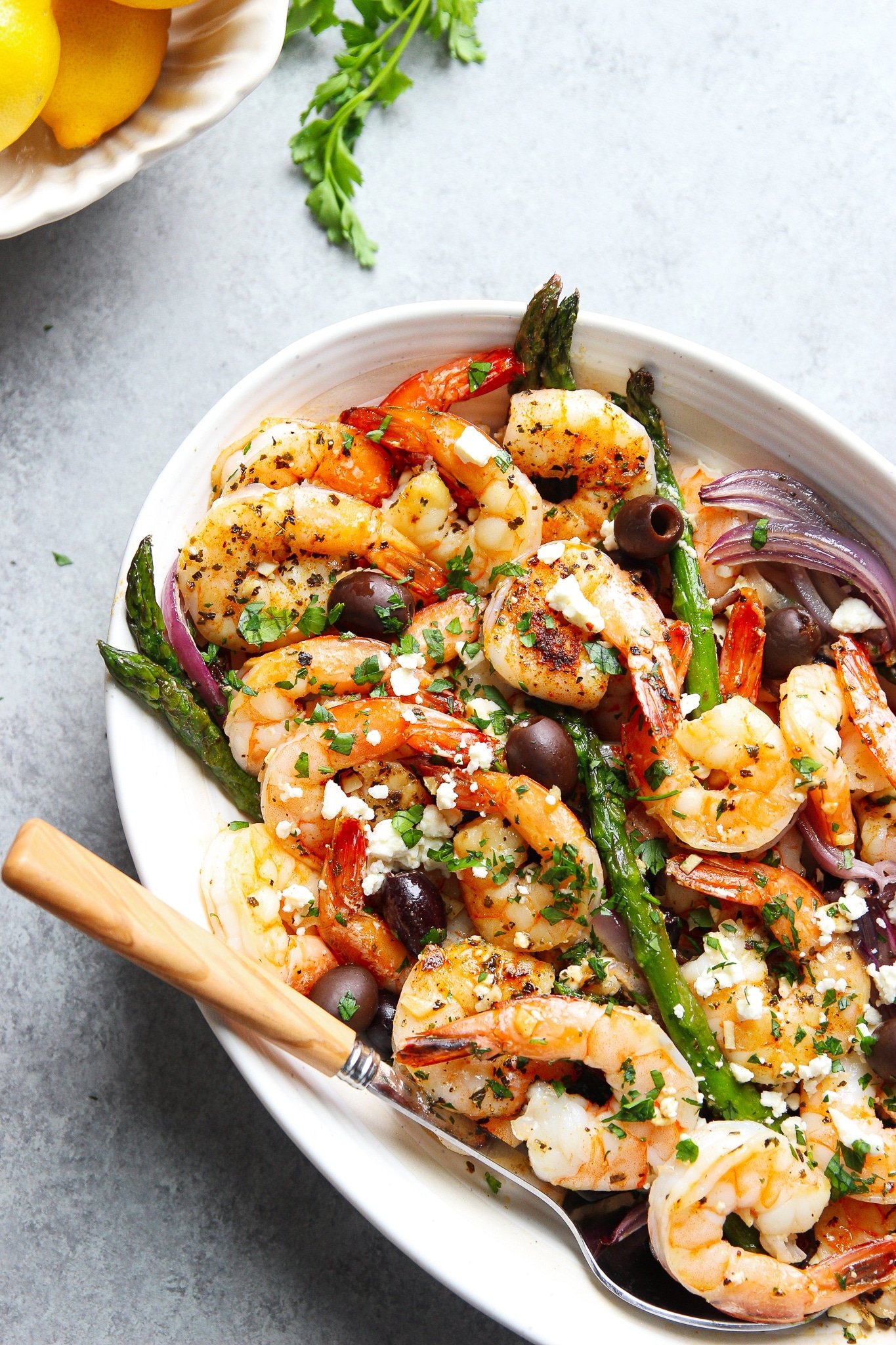 Mediterranean shrimp recipe with asparagus, onions, black olives, and feta cheese served in a white oval plate with a spoon