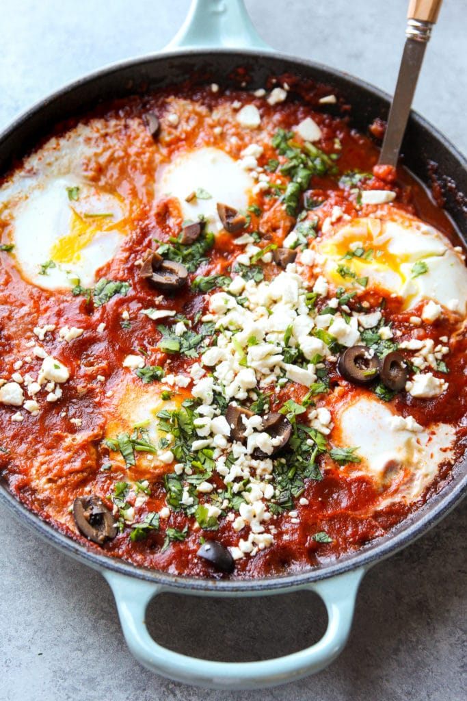 Shakshuka topped with black olives, parsley and feta cheese in a skillet with a big spoon