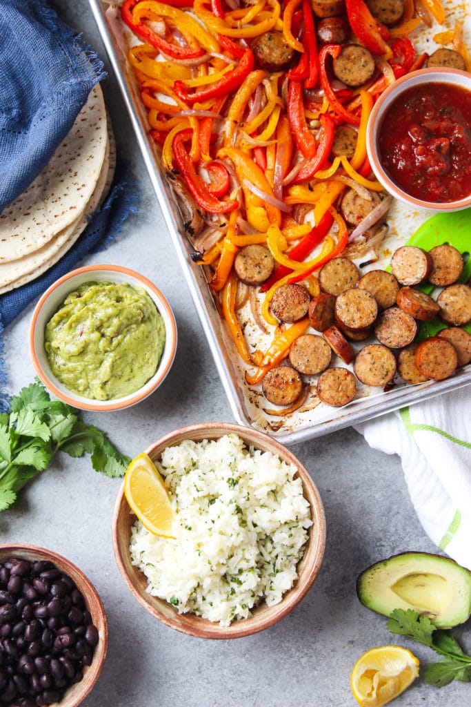 A sheet pan chicken fajitas surrounded by flat bread, a bowl of guacamole, a bowl or rice, a bowl of black beans, half a slice of avocado and some parsley