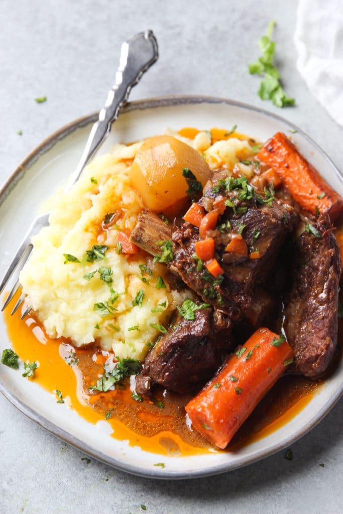 beef stew with short ribs, potatoes and carrots served over mashed potatoes on a round grey plate and a fork