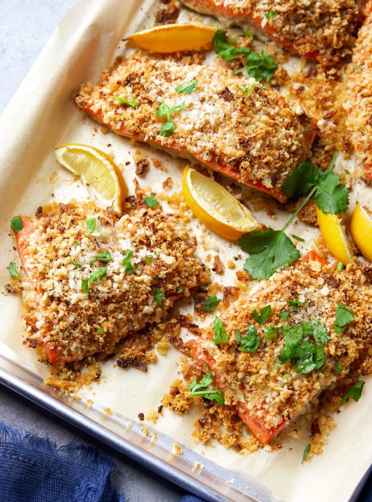 A tray lined with parchment paper contains lemon slices and crispy salmon topped with toasted onions, chopped parsley, and Parmesan