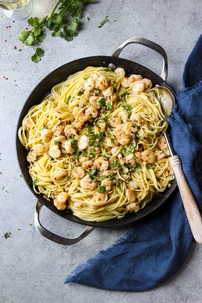 Shrimp scampi pasta in a skillet and a large spoon with a wooden handle surrounded by fresh cilantro and a blue kitchen cloth