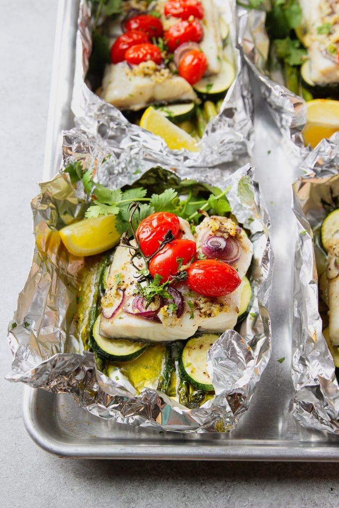 fish baked in foil with asparagus and zucchini, topped with red grape tomatoes and red onion slices. Garnished with fresh cilantro and lemon slice. Served on a sheet pan