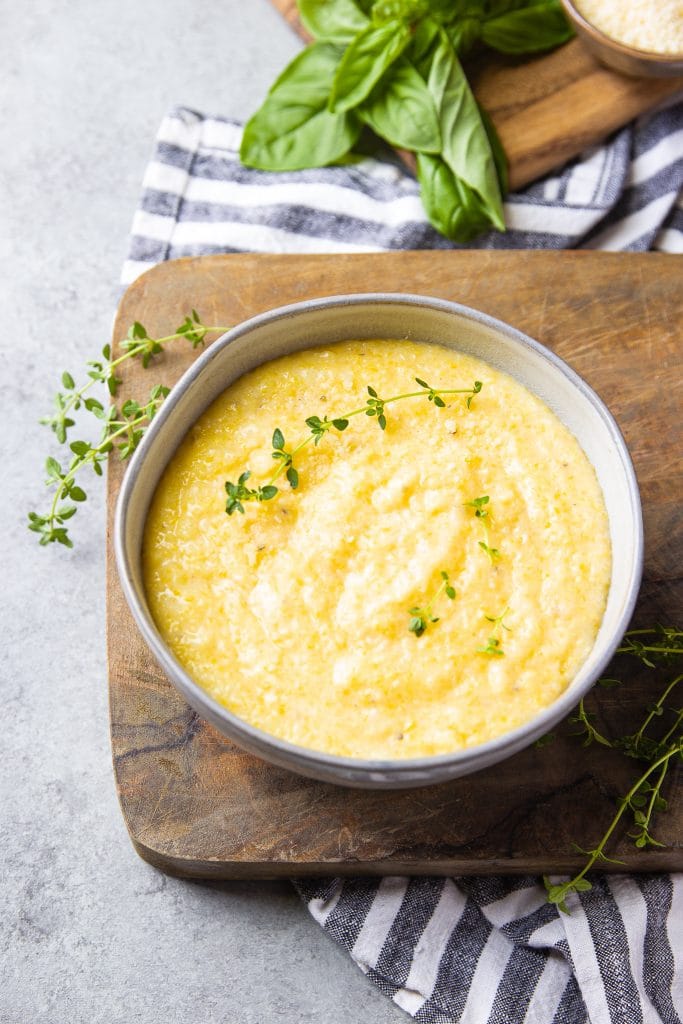 creamy polenta in a bowl sitting on a wooden board with fresh herbs