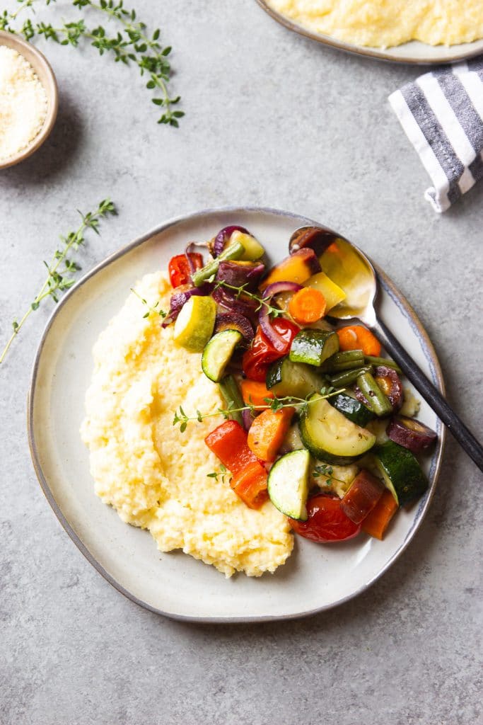 Roasted Vegetables zucchini, peppers, tomatoes over creamy polenta on a round plate with silver spoon