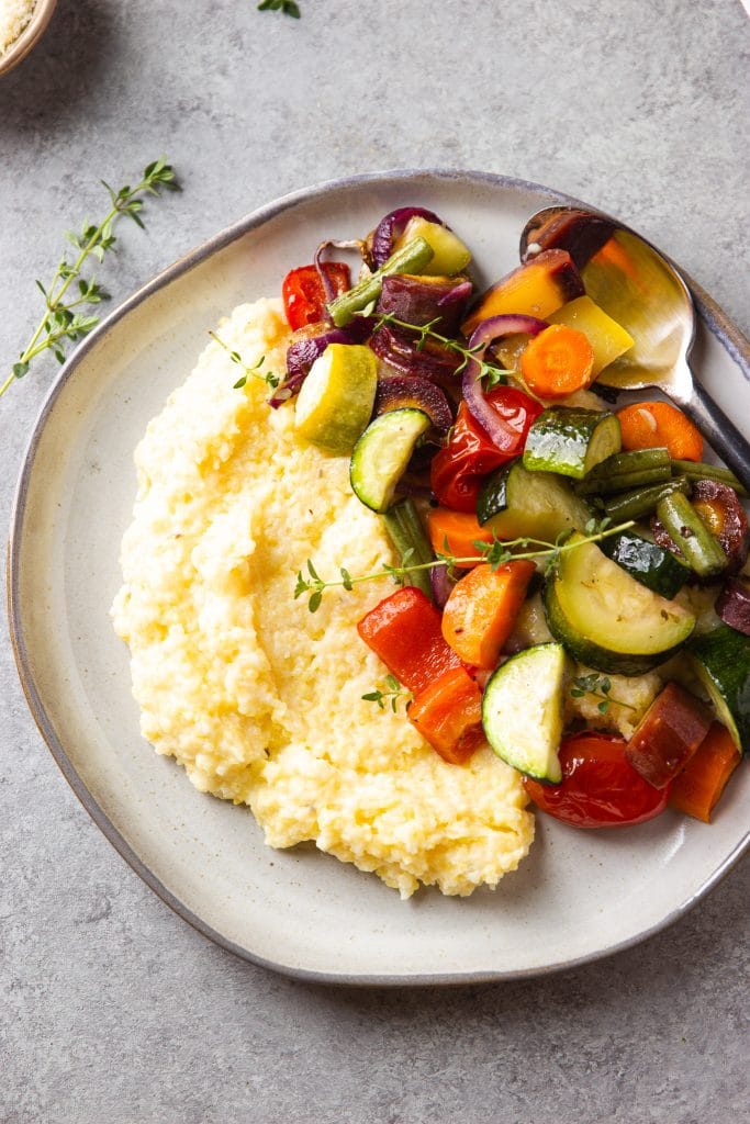 Roasted Vegetables zucchini, peppers, tomatoes over creamy polenta on a round plate with silver spoon