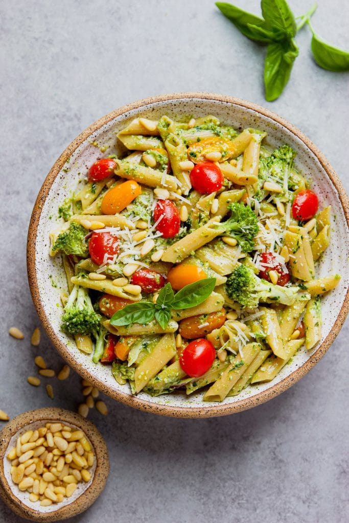 large round bowl with pesto pasta salad, fresh tomatoes, broccoli and small bowl of pine nuts.