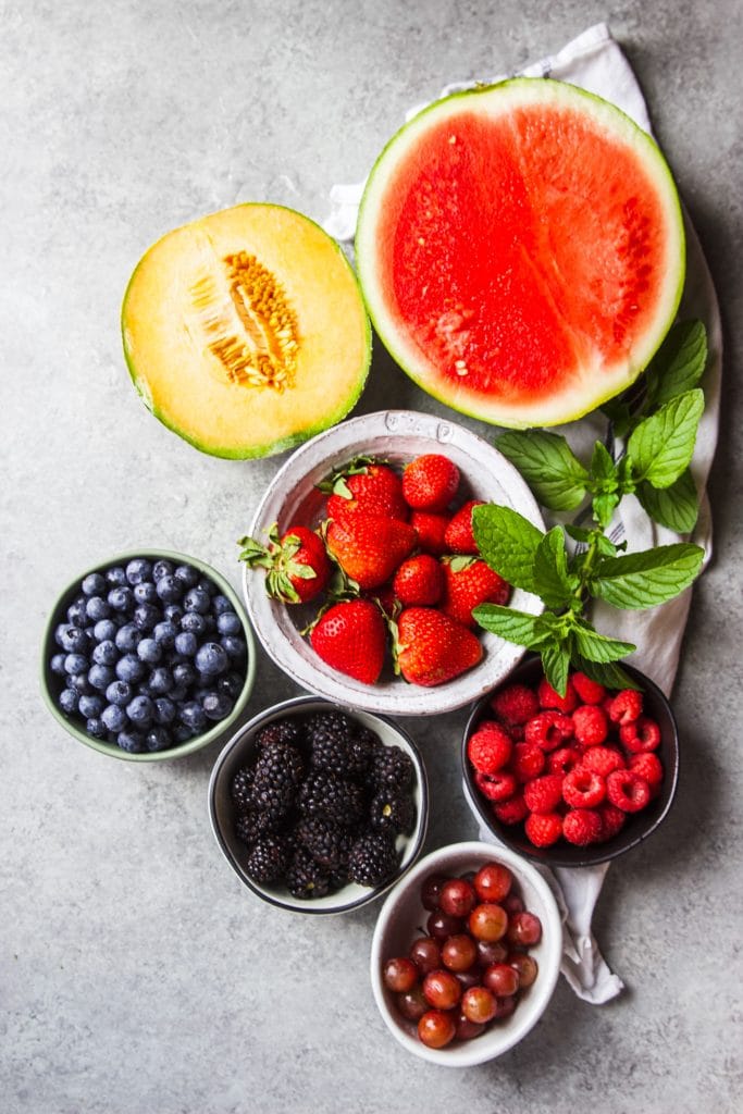 watermelon and cantaloupe sliced in half. Strawberries, blueberries, raspberries, blackberries and grapes in round bowls. Mint leaves and kitchen towel.  