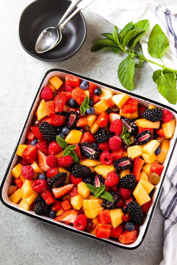watermelon, berries, cantaloupe fruit salad in square dish with mint leaves. black round serving bowl with two spoons.