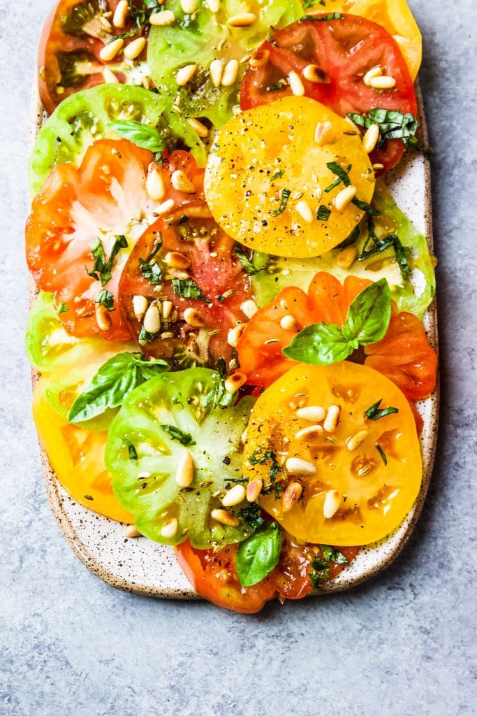 Green, yellow, orange and red slices of heirloom tomatoes on a rectangular platter. Garnished with pine nuts and basil.