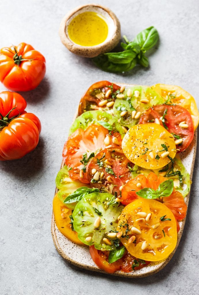 Green, yellow, orange and red slices of heirloom tomatoes on a rectangular platter. Garnished with pine nuts and basil. A round vessel with olive oil.