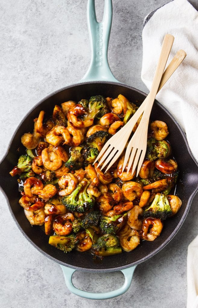 shrimp stir fry with broccoli served in a light blue Le Creuset cast iron skillet, with two wooden spoons and kitchen towel 
