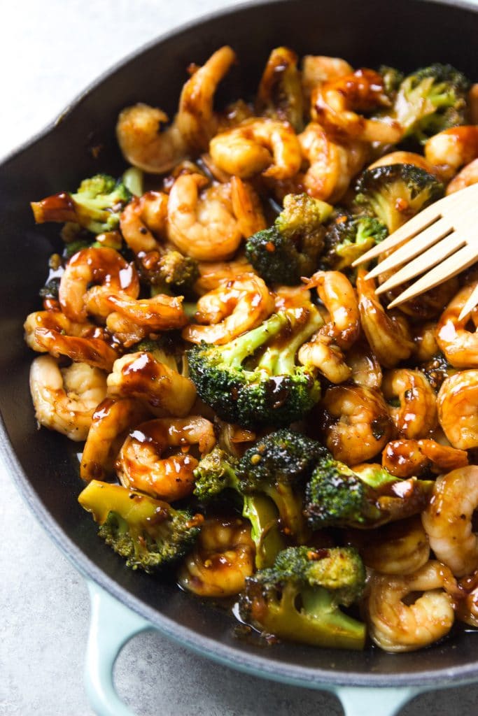 shrimp stir fry with broccoli served in a light blue Le Creuset cast iron skillet, with two wooden spoons