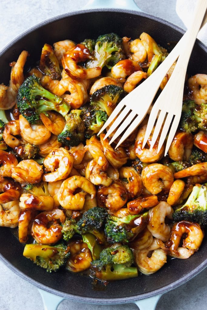 shrimp stir fry with broccoli served in a light blue Le Creuset cast iron skillet, with two wooden spoons.