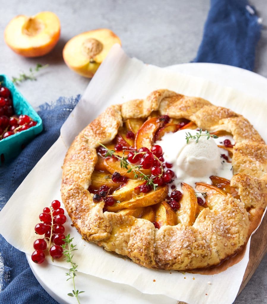 Rustic peach galette with homemade buttery crust, juicy peaches and fresh currants, a scoop of vanilla ice cream, sitting on parchment paper