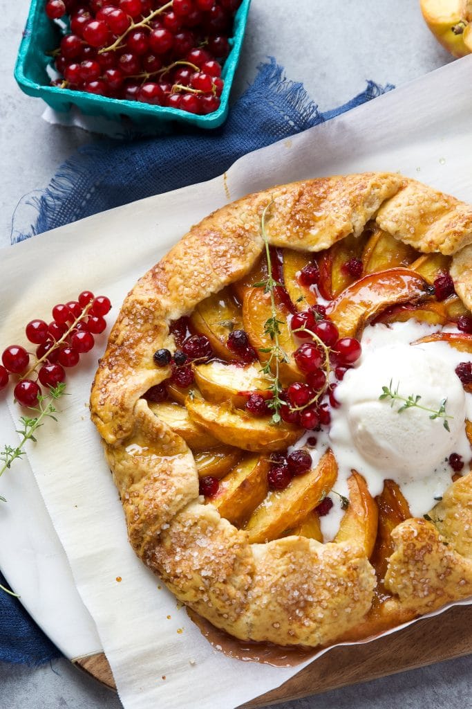Rustic peach galette with homemade buttery crust, juicy peaches, fresh currants and vanilla ice cream, sitting on parchment paper