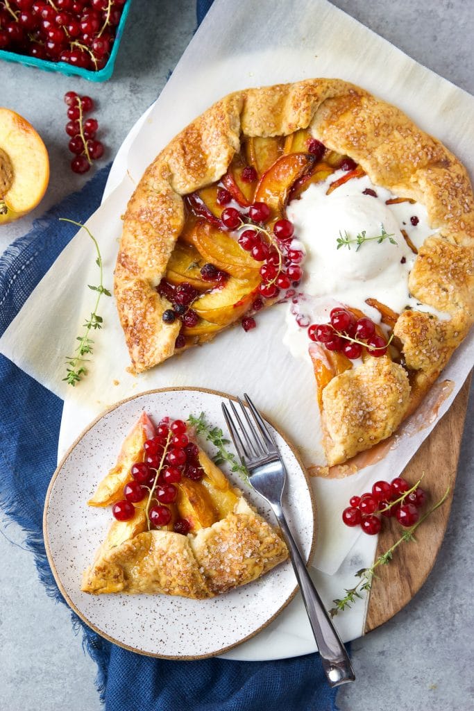 Rustic peach galette with homemade buttery crust. A slice of peach galette on a plate. Fresh currants on top and vanilla ice cream