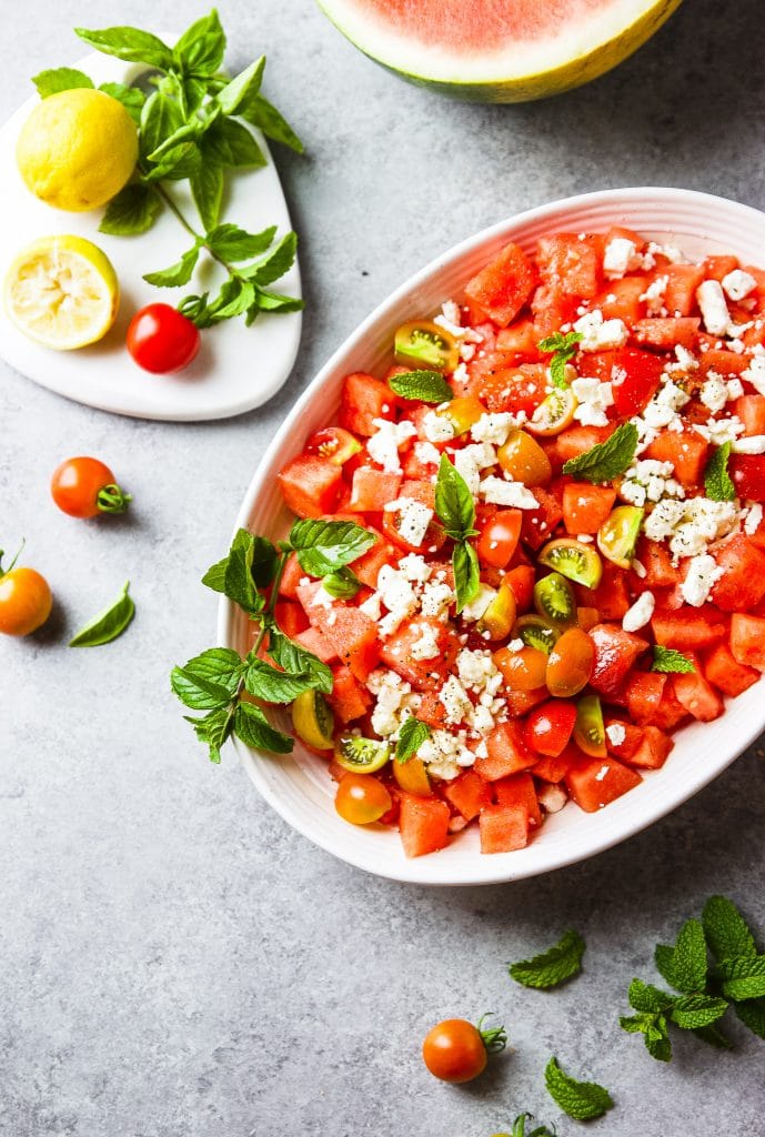watermelon feta salad with fresh mint and basil leaves. garden tomatoes. served in large white oval bowl. fresh leaves around the table