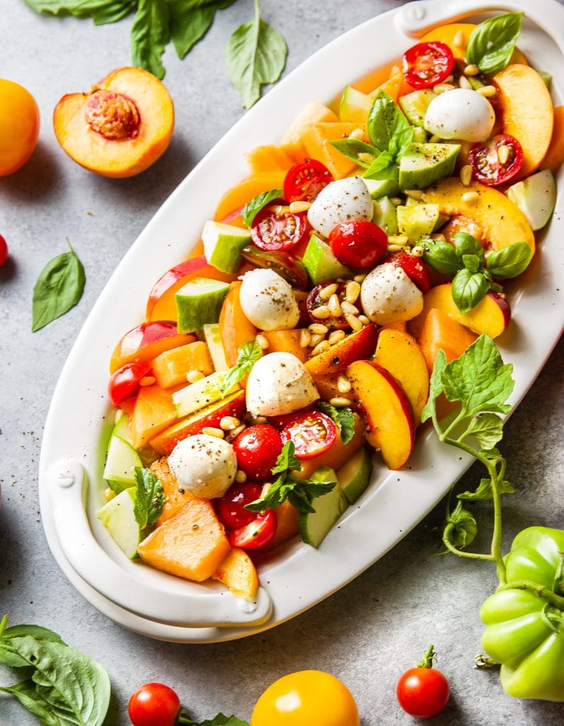 fruit salad in a white oval platter with cut up cantaloupe, peach slices, cherry tomatoes, cucumber slices and fresh mozzarella. Topped with basil and mint leaves. 