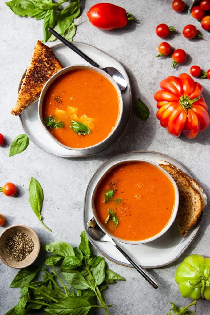 two bowls of tomato soup with spoons and toast. Fresh tomatoes and basil displayed