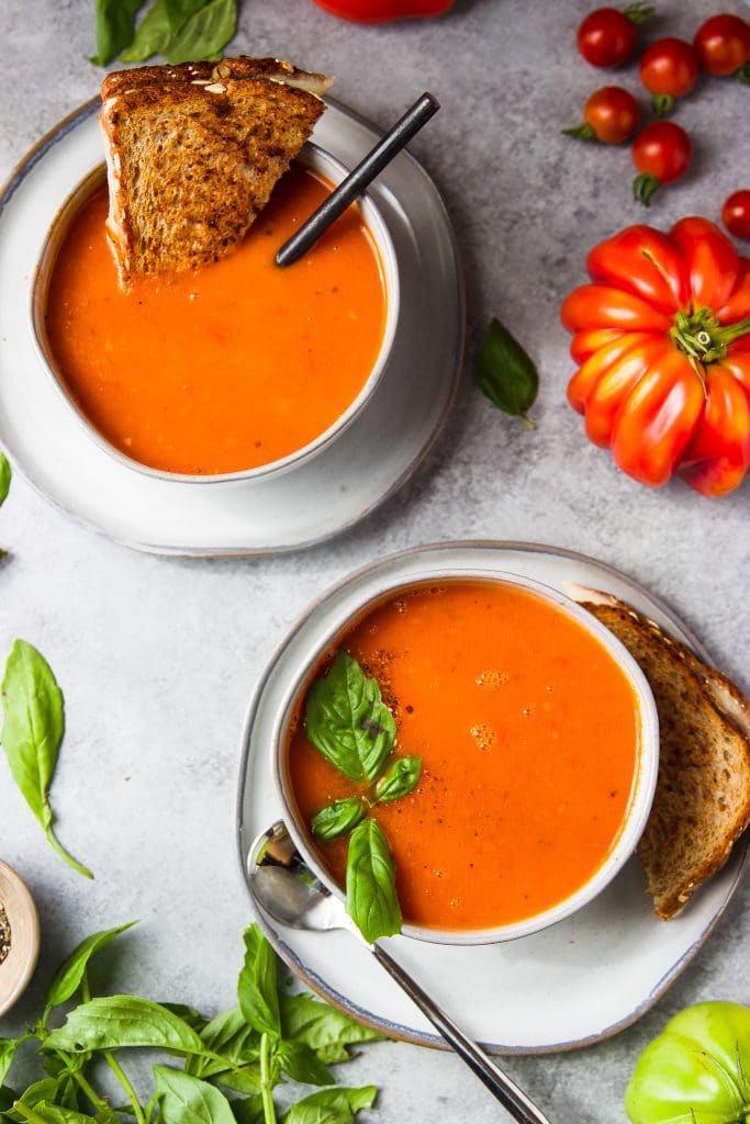 two bowls of tomato soup with spoons and toast. Fresh tomatoes and basil displayed