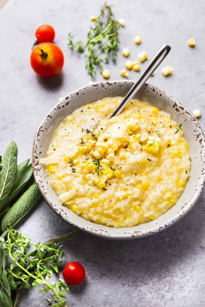 creamy corn risotto in a bowl with grated parmesan cheese, fresh thyme and a spoon. Fresh herbs and veggies on the table.