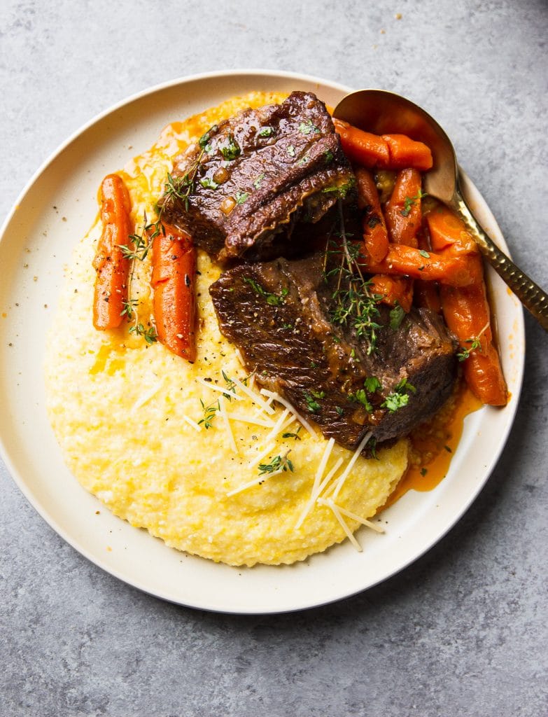 A round plate with instant pot beef short ribs, carrots and creamy polenta. Fresh herbs as garnishing and parmesan cheese. 