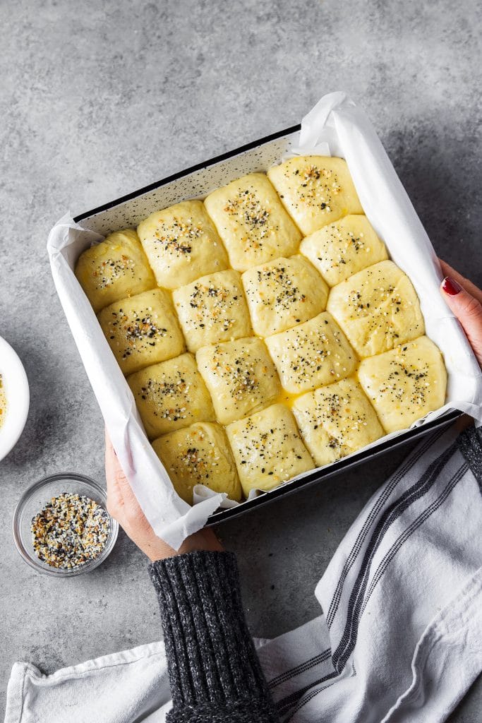 A hand holding a baking dish with dinner rolls ready to go in the oven. A small bowl with seasoning