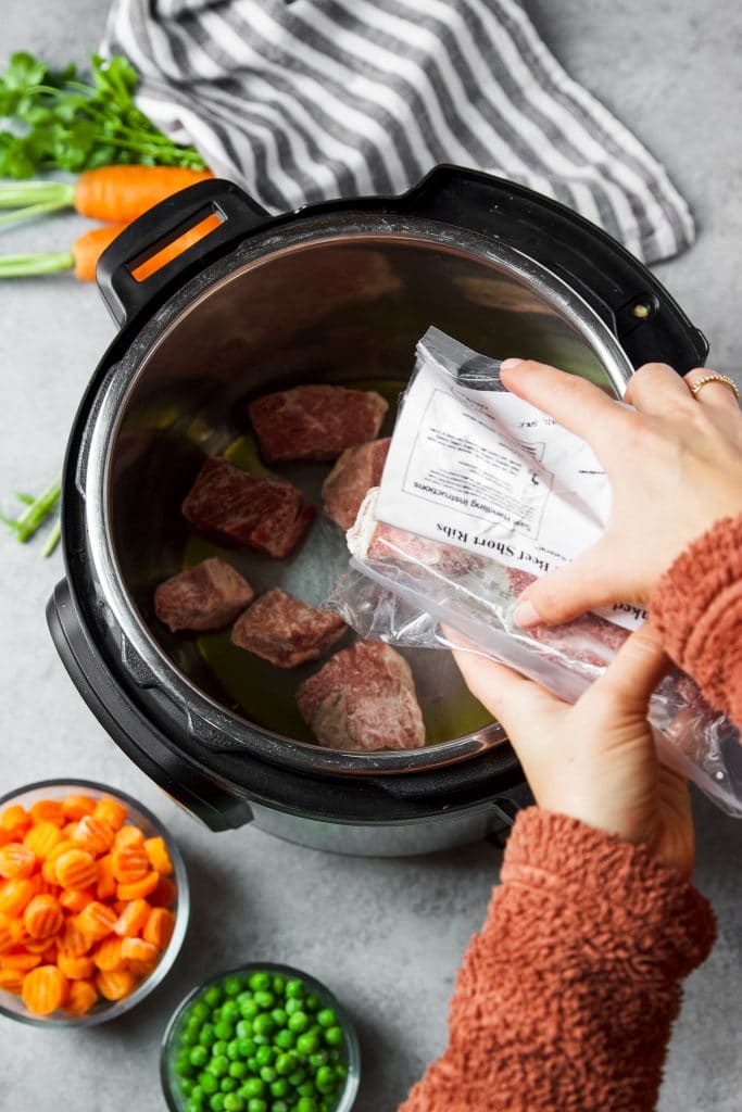 beef stew being added to the instant pot. A bowl of sliced carrots and a bowl of fresh peas.