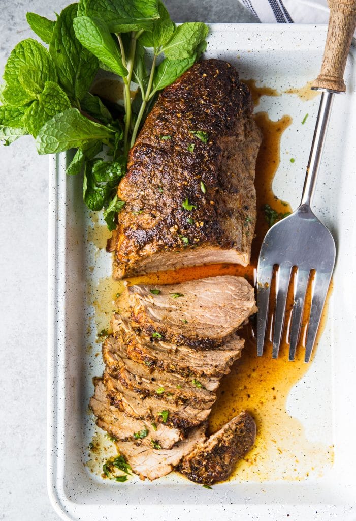 Sliced pork tenderloin in a baking pan seasoned with herbs. A large serving fork next to the pork.